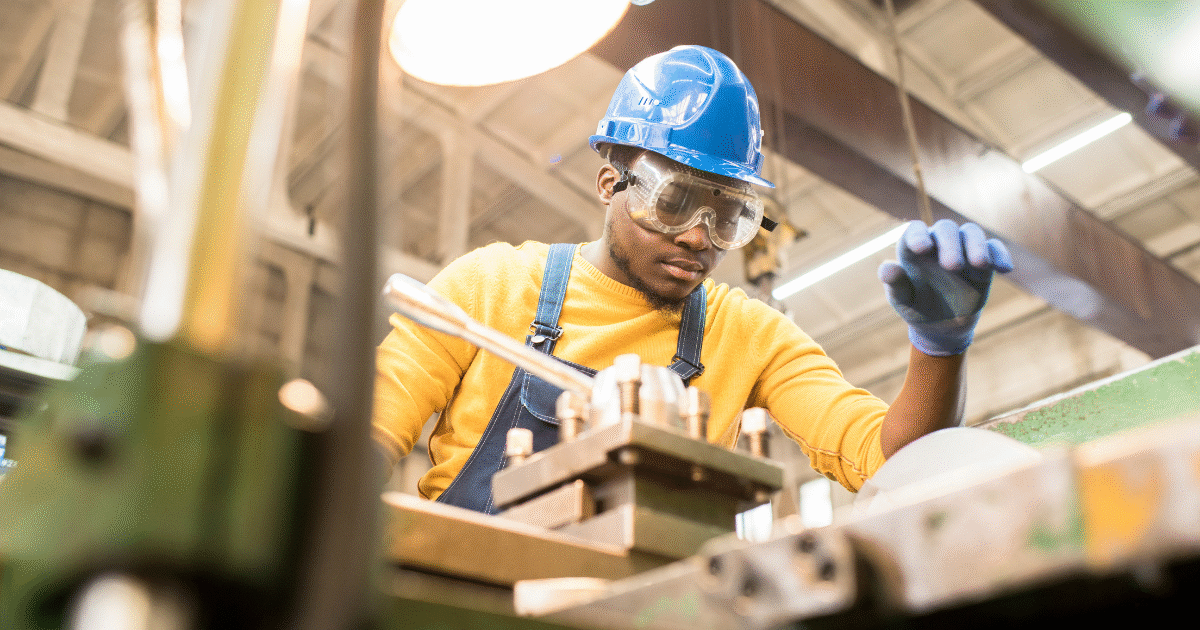 Serious young black factory engineer in hardhat and safety goggles examining milling lathe and repairing it while working at a production plant