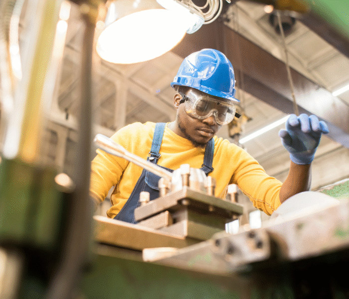 Serious young black factory engineer in hardhat and safety goggles examining milling lathe and repairing it while working at a production plant