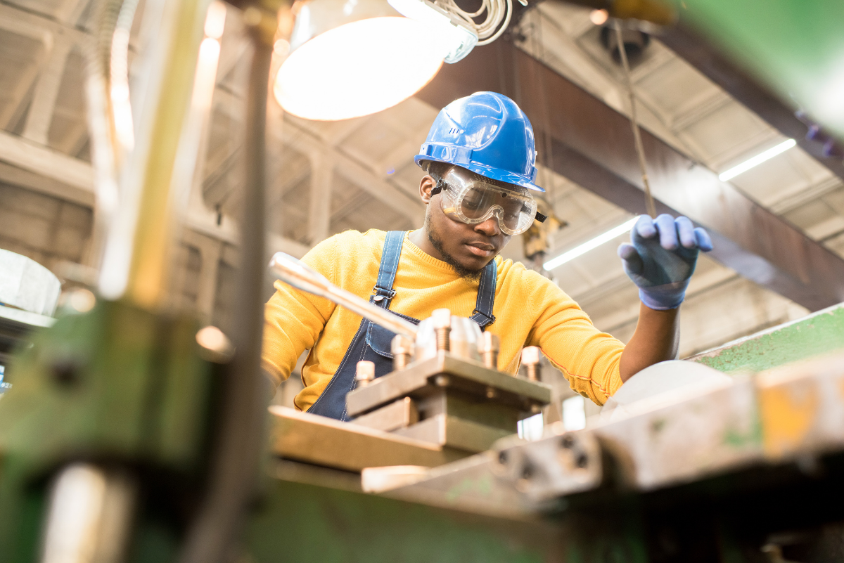 Serious young black factory engineer in hardhat and safety goggles examining milling lathe and repairing it while working at a production plant